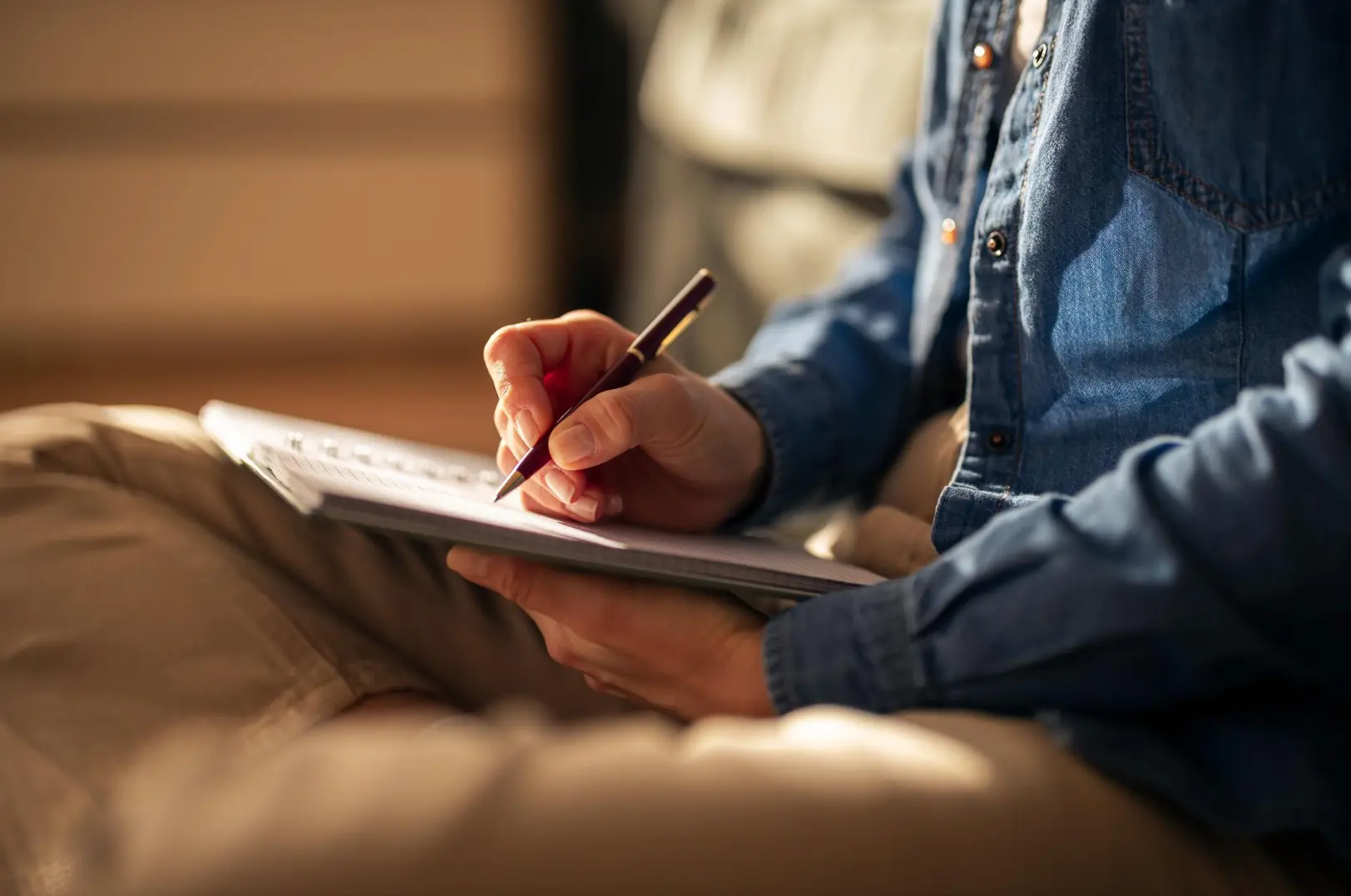 Person in denim shirt journaling with a pen in warm light